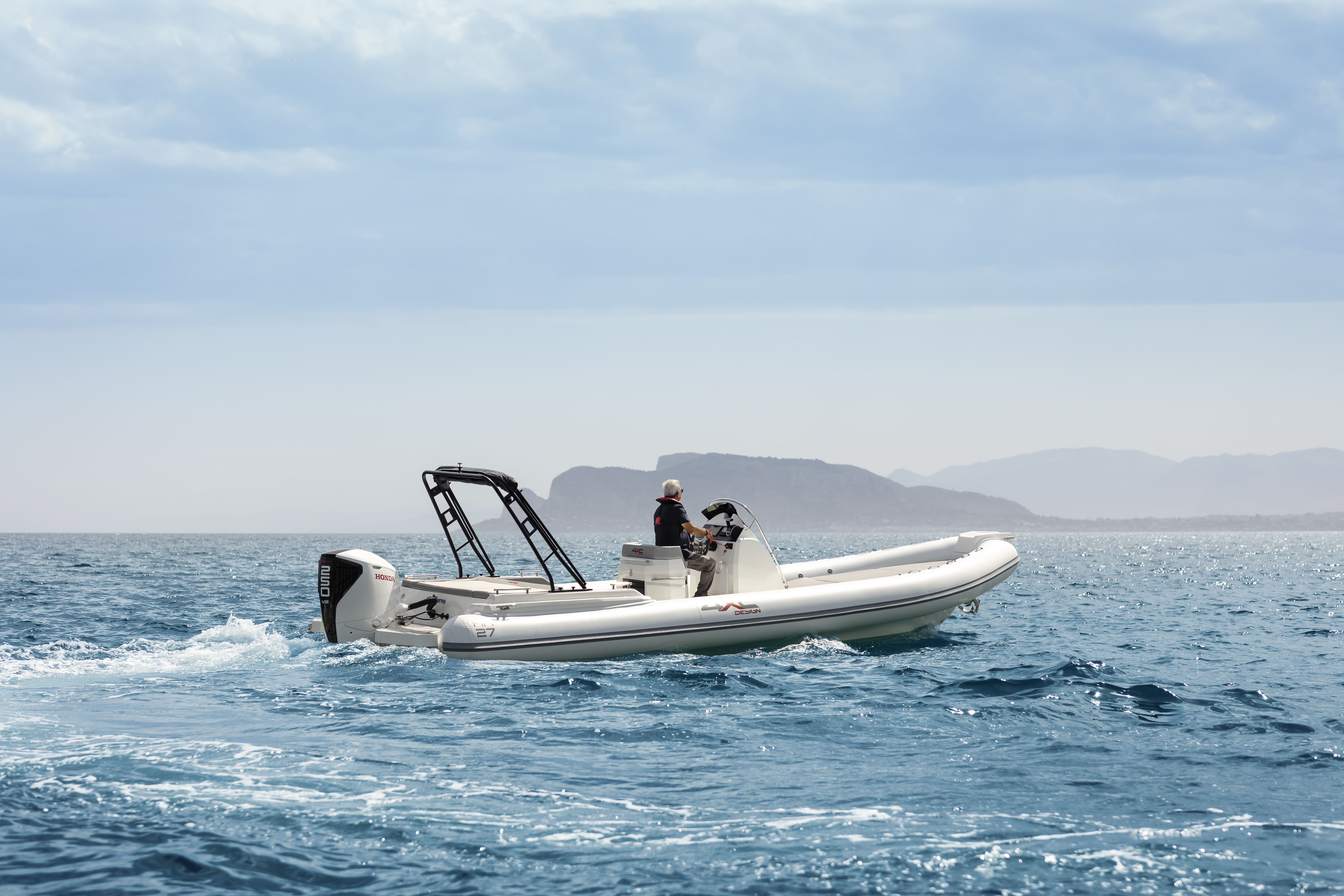 A white Honda inflatable boat with an outboard motor speeds across calm blue waters. Mountains are visible in the distance under an overcast sky.